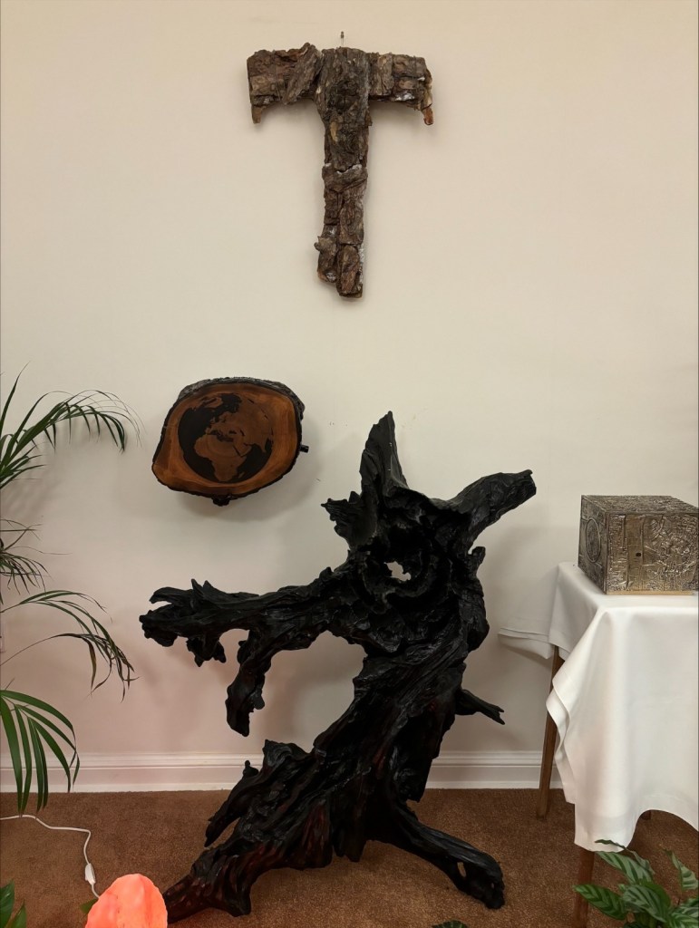 Image of the Tabernacle on wall bracket. Above it is a cross made from bark, below it a dark wooden sculpture, and o the left a silver box is on a table.